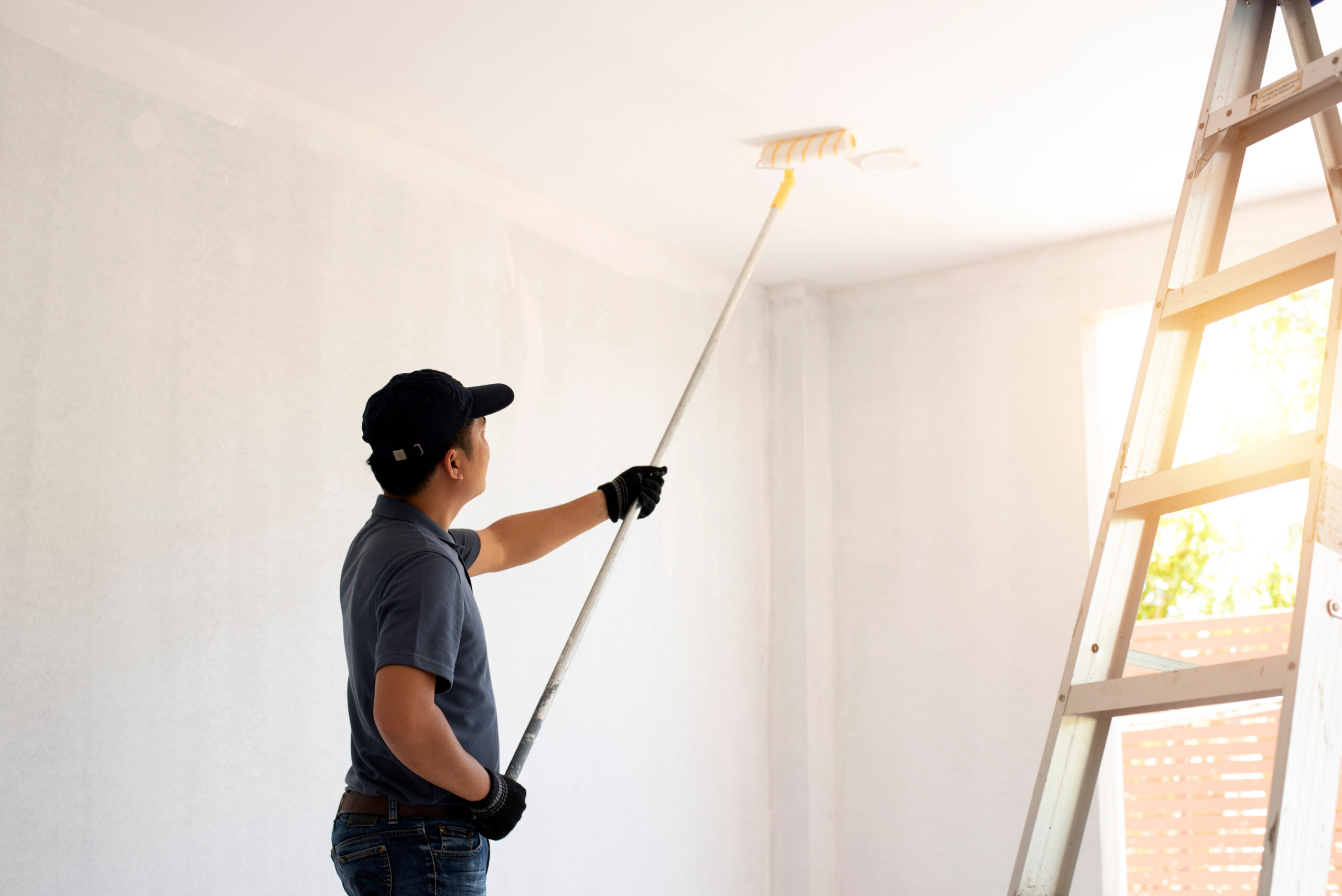 Asian rear view of a male painter drawing a wall with paint roller and a separate tank from a large empty space with wooden stairs.