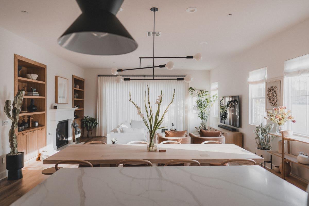 Modern open-concept living and dining area with a marble kitchen island, wooden dining table, white sofa, and indoor plants, lit by natural light from large windows.