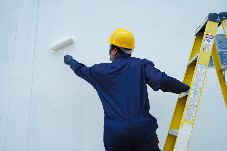 Worker in a yellow hard hat and navy uniform painting a white wall with a roller next to a yellow ladder.