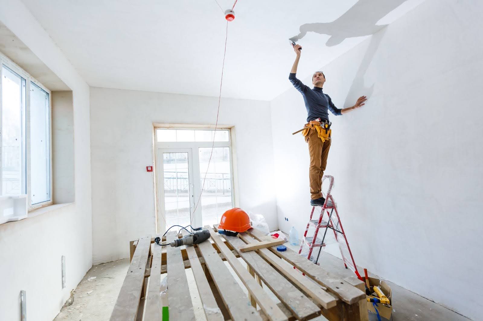 Painter standing on a ladder atop a workbench, painting high ceiling in a home renovation project.